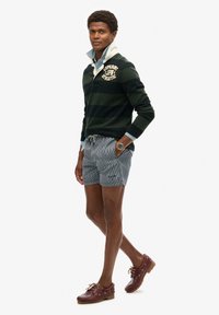 Suéter de rayas verde oscuro con cuello, pantalones cortos azul grisáceo con rayas verticales y zapatos de barco granate. Acentuado con un reloj de pulsera.
