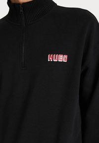 Felpa nera con zip a quarti, colletto a costine e logo "HUGO" ricamato in rosso e bianco sul lato sinistro del petto.
