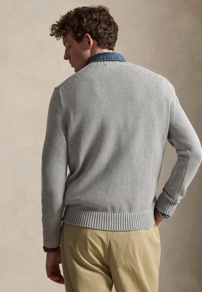Man met krullend haar die een lichtgrijze trui draagt over een blauw overhemd met kraag en beige broek, staand met zijn rug naar de kijker gekeerd.
