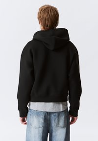 Črna obrezana majica s kapuco iz mehke tkanine z veliko kapuco in rebrastimi manšetami, nošena preko svetlo sive majice ter kombinirana s svetlo modrimi jeans hlačami.