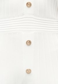 Λευκό ριμπ ύφασμα με οριζόντια και κάθετα υφαντά σχέδια, με τρία στρογγυλά χρυσά κουμπιά με περίπλοκα μοτίβα.