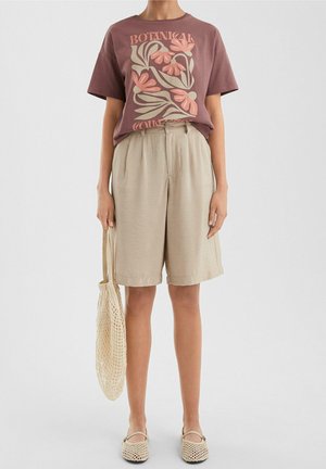 Person iført en mauve t-shirt med blomstermotiv, beige højtaljede shorts, vævede beige sko og holder en beige netpose.