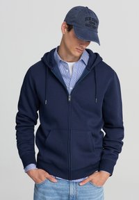 Felpa navy con zip e tasca a marsupio, realizzata in tessuto morbido; indossata sopra una camicia a righe blu chiaro, abbinata a jeans blu chiaro.