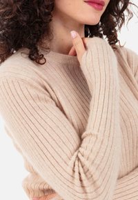 Beiger, gerippter Pullover mit langen Ärmeln, rundem Ausschnitt und weicher Textur, mit markanten Daumenloch-Details an den Ärmelenden.