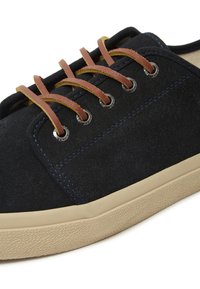 Crni nubuk tenisice s bež gumom na donjem dijelu i teksturiranim uzorkom. Ima smeđe kožne vezice i metalne oči na gornjem dijelu.