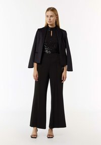 Schwarzes Paillettenoberteil mit einem Schlüsselloch-Ausschnitt, kombiniert mit einem schwarzen taillierten Blazer und weit geschnittenen Hosen, ergänzt durch schwarze Riemchensandalen.