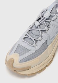 Chaussure de sport grise avec un dessus en matériaux mixtes, incluant du maille et des accents synthétiques, une semelle épaisse et un système de laçage complexe.