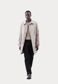 Světle béžový trench coat, zapnutý, s límcem; nošený přes světle šedý svetr a černé kalhoty; model stojí na bílém pozadí.