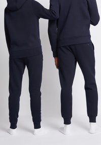 Completo con felpa e pantaloni da jogging blu navy, caratterizzato da una texture liscia e polsini a costine, indossato con calzini bianchi. Vista posteriore di due figure.