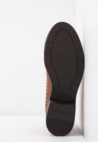 Semelle d'une chaussure en cuir marron reposant contre un mur blanc, montrant une semelle en caoutchouc noire avec la marque "Carmela" et la taille 37.
