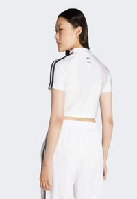 Biele skrátené športové tričko s krátkymi rukávmi, s čiernymi pruhmi a logom na zadnej strane. Vybavené bielymi nohavicami, zdôrazňujúcimi atletický dizajn.