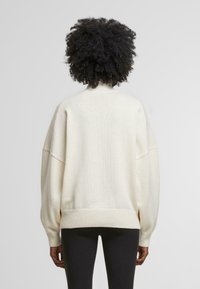 Cream-colored, oversized knit sweater with drop shoulders, ribbed cuffs and hem, and a textured finish. Back view shows a smooth, simple design.