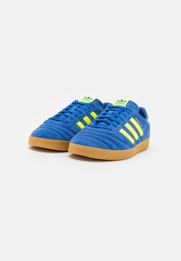 Niebieskie zamszowe sneakersy z trzema żółtymi paskami, teksturowaną gumową podeszwą i wyściełanym kołnierzem dla komfortu. Projekt zawiera akcenty z logo.