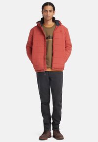 Rode gewatteerde jas met marineblauwe capuchon, gedragen over een bruine sweatshirt. Zwarte jeans en bruine laarzen maken de look compleet. Rechtdoordefrontalenzicht.