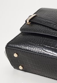 Schwarze Handtasche aus Kunstleder mit strukturiertem Krokodilmuster, goldfarbener Hardware und Schutzfüßen an der Unterseite. Magnetverschluss.