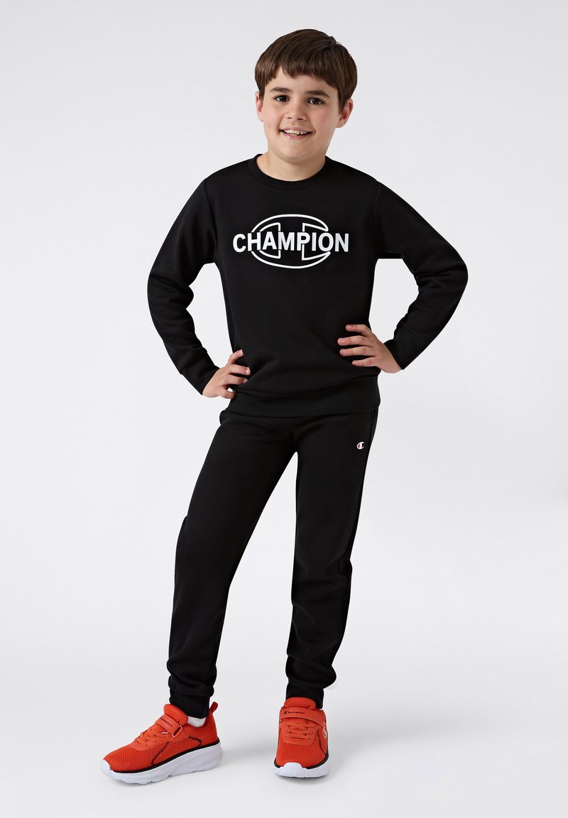 Sudadera negra con el logo "CHAMPION", pantalones de chándal negros a juego y zapatillas deportivas naranjas brillantes con suelas blancas. Tejido suave, corte atlético.
