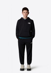 Melns sporta krekls ar kapuci un priekšējo kabatu, uz tā ir balts logotips. Kopā valkā melnas bikses un pelēkas kurpes ar tumšām zolēm.