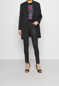 Zwarte gewatteerde blazer over een graphic T-shirt met roze en blauwe accenten, gecombineerd met glanzende zwarte skinny broek en zwarte stiletto sandalen.