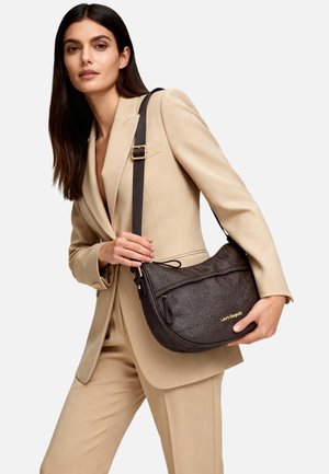 Borsa a tracolla in pelle marrone a grana con una forma curva, dettagli in oro, chiusura superiore con cerniera e tracolla regolabile, tenuta da una persona con un completo beige.