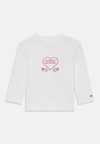 Vit bomullsskjorta med långa ärmar som har en rosa hjärtformad design med texten "TOMMY HILFIGER" och blommiga accenter. Tryckknappdetalj på axeln.