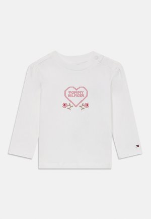 Camisa de algodón blanca de manga larga con un diseño de corazón rosa y el texto "TOMMY HILFIGER" acompañado de acentos florales. Detalle de botón a presión en el hombro.