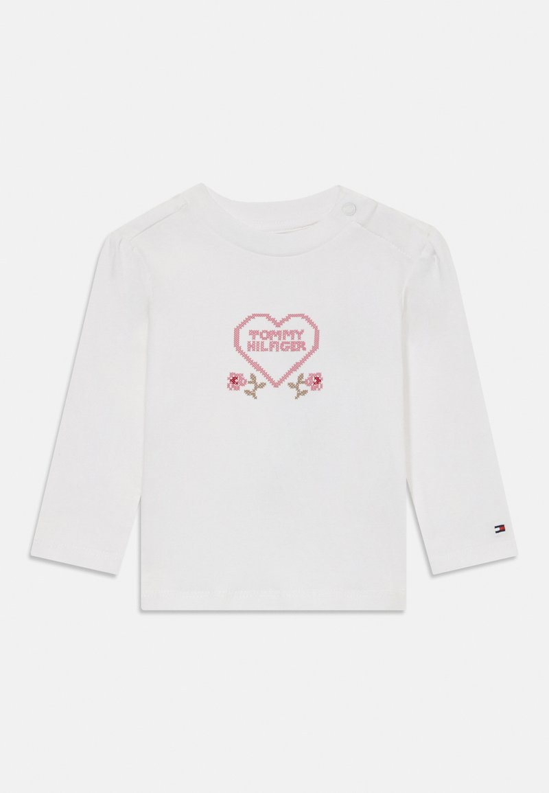 Vit bomullsskjorta med långa ärmar som har en rosa hjärtformad design med texten "TOMMY HILFIGER" och blommiga accenter. Tryckknappdetalj på axeln.