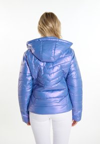 Giacca puffer viola lucida con cappuccio, caratterizzata da un design trapuntato e cuciture angolate, indossata sopra pantaloni bianchi.