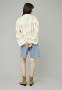 Veste claire texturée avec des motifs géométriques, associée à un short en jean et des bottines à talons marron. La vue arrière met en avant le design global.