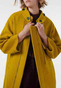 Abrigo de lana amarillo con acabado texturizado, gran cuello y botones verdes. Cuenta con una cremallera frontal y bolsillos laterales. La modelo agarra el cuello.