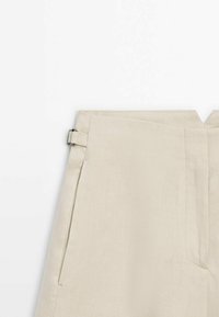 Pantalon beige taille haute avec détail de boucle latérale, coutures droites, et fermeture avant cachée, présenté sur fond blanc.