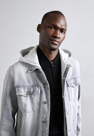 Lichtgrijze denimjack met een contrasterende grijze capuchon, voorzakken en knoopdetails, gedragen over een zwart ribbeltrui.