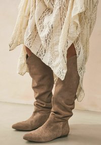 Botas marrones de ante por encima de la rodilla con un ajuste relajado, punta afilada y costuras decorativas. Combinadas con una prenda de encaje crema texturizada.