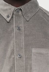 Camisa de pana gris con textura acanalada, cierre de botones en la parte delantera y un bolsillo en el pecho, que presenta cuatro botones gris oscuro y un cuello puntiagudo.