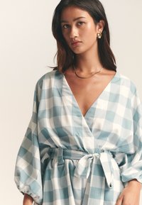 Mėlynos ir baltos spalvų languota suknelė su apvyniojamu dizainu, gilus V formos iškirptė ir pūstos rankovės. Yra derantis diržas liemeniui pabrėžti.