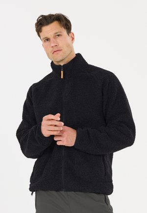 Man met kort bruin haar draagt een zwarte fleecejas met rits en staat tegen een effen witte achtergrond met de handen ineen.