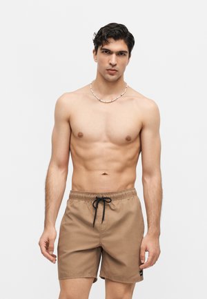 Άνδρας χωρίς μπλούζα που φορά ταμπά μαγιό με μαύρο κορδόνι και περιδέραιο με χάντρες, στέκεται μπροστά σε απλό, ανοιχτόχρωμο φόντο.