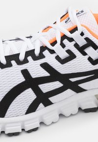 Scarpa da ginnastica bianca con una parte superiore in mesh testurizzato, accenti neri del logo, dettagli arancioni vivaci e una suola bianca spessa con aderenza.