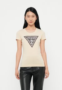 Camiseta de manga corta color beige claro con un logo triangular en negro y plata que presenta el texto "GUESS?" y acentos decorativos en el centro.