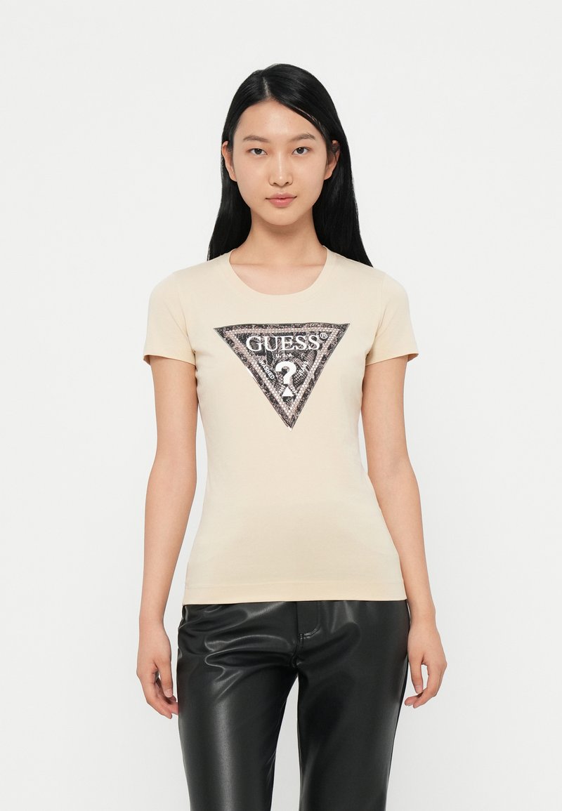 Camiseta de manga corta color beige claro con un logo triangular en negro y plata que presenta el texto "GUESS?" y acentos decorativos en el centro.