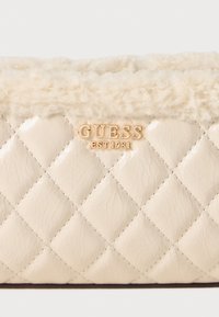 Gequilted crèmekleurige tas met een zachte nepbont afwerking, voorzien van gouden "GUESS" branding en een gestructureerde afwerking.