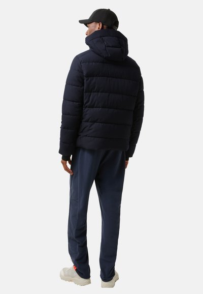 Námořnická puffer bunda s kapucí, která má horizontálně prošívané panely a elastické manžety, v kombinaci s odpovídajícími námořnickými tepláky a světlými teniskami.