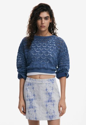 Blauer strukturierter, verkürzter Pullover mit einem Rautenmuster und weiten Ärmeln, kombiniert mit einem hellgrauen Rock, der blaue abstrakte Flecken aufweist.