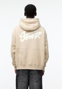 Helle beiger Kapuzenpullover mit einem großen weißen Grafiklogo auf dem Rücken. Hergestellt aus weichem Material, mit Kapuze und lässiger Passform.