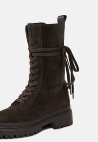 Hnědé semišové ботy до kotníků na šněrování s chunky podešví, kulatou špičkou a stuhovými šňůrkami svázanými vzadu pro přidaný detail.