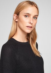 Pull en maille noire avec une texture côtelée, encolure ronde et manches longues. Finitions délicates le long des épaules et silhouette ajustée.