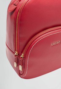 Zaino in pelle rossa con finitura testurizzata, caratterizzato da accessori in oro, doppie cerniere e cuciture bianche lungo il design curvato.