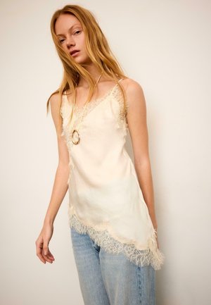 Jeune femme en haut crème sans manches orné de dentelle et jean bleu clair, portant un long collier pendentif, debout devant un fond uni.