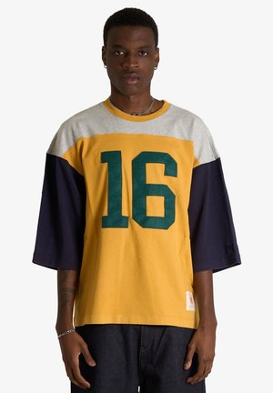 Giovane uomo che indossa una maglietta gialla, grigia e blu navy con il numero 16 davanti, in piedi davanti a uno sfondo bianco semplice.