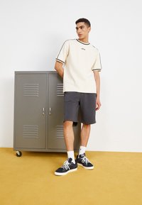T-shirt crema a maniche corte con bordi neri, pantaloni corti grigi e sneaker nere. Armadietto grigio sullo sfondo su un pavimento giallo.