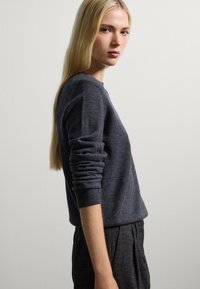 Pull en tricot gris foncé avec des manches longues, un col rond et des poignets côtelés, associé à un pantalon texturé foncé.